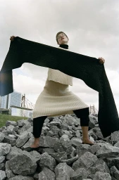 Model standing on a rocky terrain holding a long black scarf horizontally, wearing a cream knit set against an overcast sky.