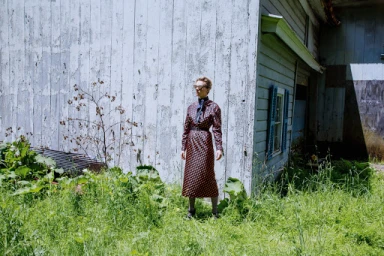Model standing outdoors in a patterned long-sleeve dress before a weathered white wooden building and tall overgrown grass.