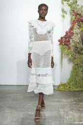 Model in a white crocheted lace dress standing on a concrete floor against a white wall with floral installation