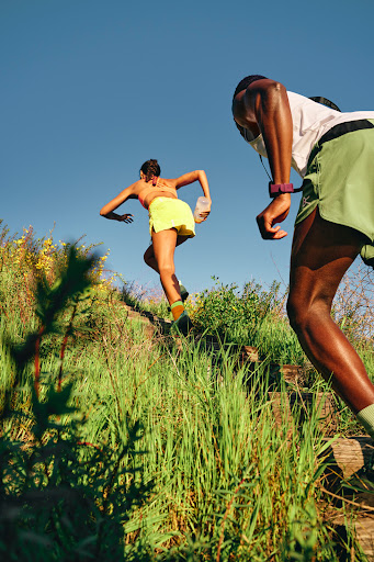 Two athletes run uphill on a grassy trail under a clear blue sky, one Black male in the foreground and a woman in the background.