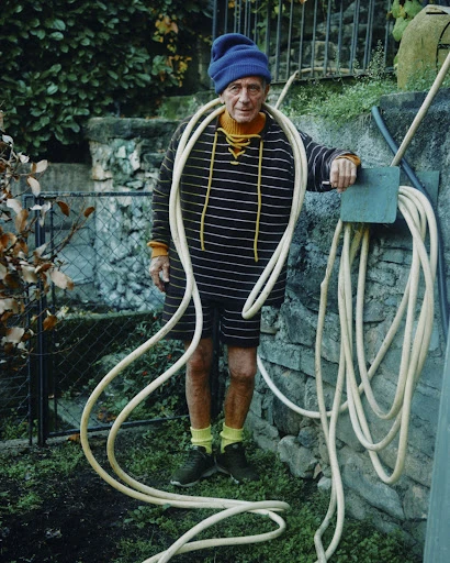Elderly adult male in outdoor portrait. Wearing knit hat and striped shirt, posing with coiled cream-colored garden hose and outdoor wall.