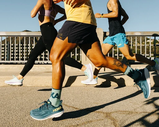 Three athletic people are captured mid-run on a paved road, showcasing athletic apparel and Hoka branded running shoes against blue sky.