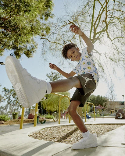 Young boy outdoors in park wearing Air Jordan shorts and sneakers poses with a kick while barefoot and tree branches overhead.