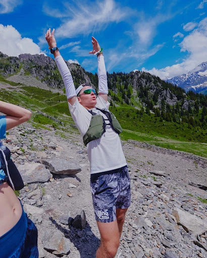 Adult male trail runner stretching arms overhead in a mountain landscape with rocks and grass against a partially cloudy blue sky.