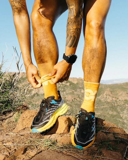 Athlete laces up running shoes outdoors. Focus on running shoes, socks, and tattoos in a mountain landscape setting on a sunny day.