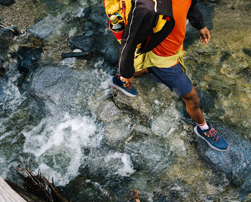 Person hiking across a rocky stream with clear water, wearing orange shirt, blue shorts, backpack, and hiking shoes, top-down perspective.