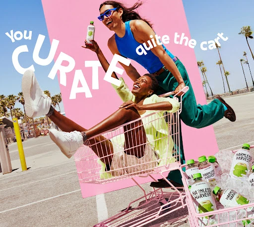 Two women laugh while riding in a shopping cart full of bottled drinks against a backdrop of palm trees and a bright sky.