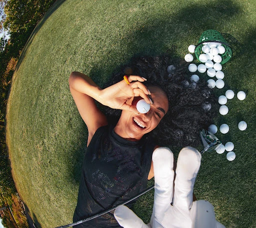 Woman lying on grass holding golf ball, smiling in wide shot with golf club and golf balls, happy and enjoying the sport.