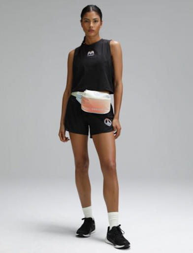 Athletic woman stands in a studio wearing a black tank top and shorts with black sneakers, white socks and a fanny pack.