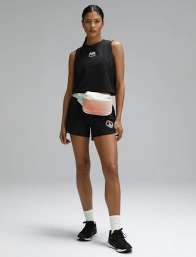 Athletic woman stands in a studio wearing a black tank top and shorts with black sneakers, white socks and a fanny pack.