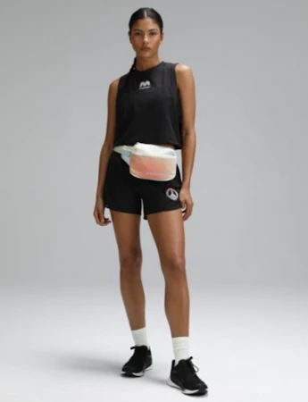 Athletic woman stands in a studio wearing a black tank top and shorts with black sneakers, white socks and a fanny pack.