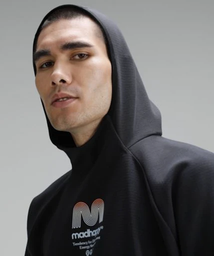 Close-up of an adult male wearing a hooded black sweatshirt with logo detail against a plain gray studio backdrop. Fashion shot.