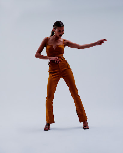 A woman poses in a warm brown two-piece outfit including a crop top and high-waisted pants with heeled sandals on a neutral background.