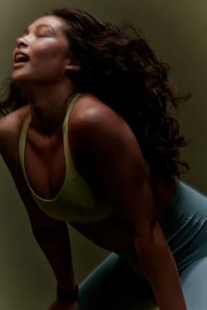 Profile shot of a woman with curly hair wearing a sports bra and leggings. She is backlit, with her head tilted back.