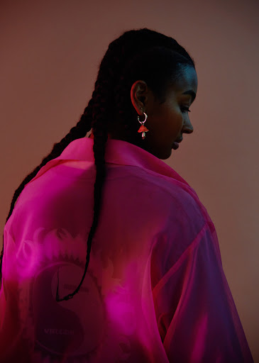 A young Black woman in a pink sheer top poses, her braided hair cascading down her back, against a soft-toned gradient background.