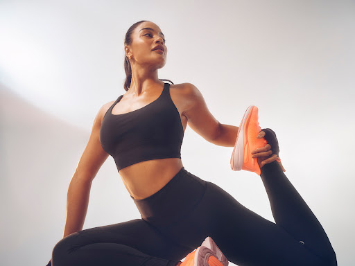 An adult female in black activewear poses from a low angle, holding her foot with one hand, wearing neon orange athletic shoes.