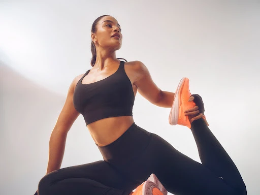 An adult female in black activewear poses from a low angle, holding her foot with one hand, wearing neon orange athletic shoes.