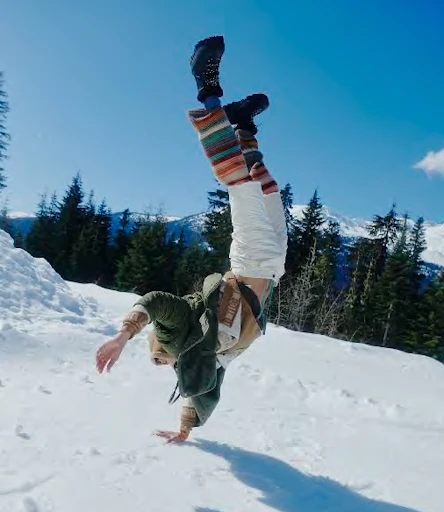 A person does a handstand in the snow wearing colorful socks, white pants, boots, and an olive-green jacket on a sunny day.