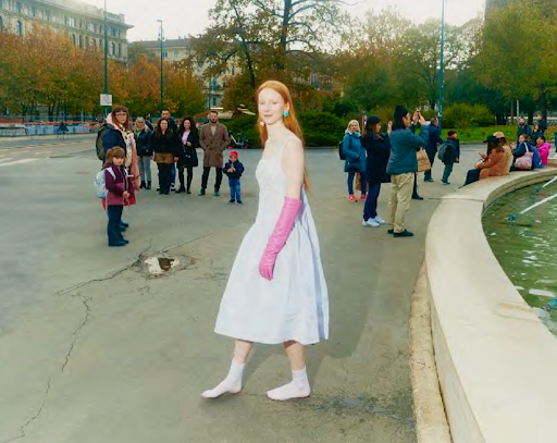 Red-haired woman in a white dress and pink gloves walks across a grey plaza with other people in the background on a sunny day.