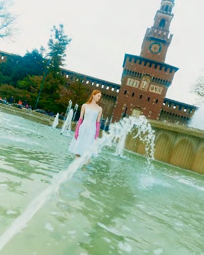 A fair-skinned adult woman in a white dress and pink gloves poses in a fountain with a brick tower and people in the background.