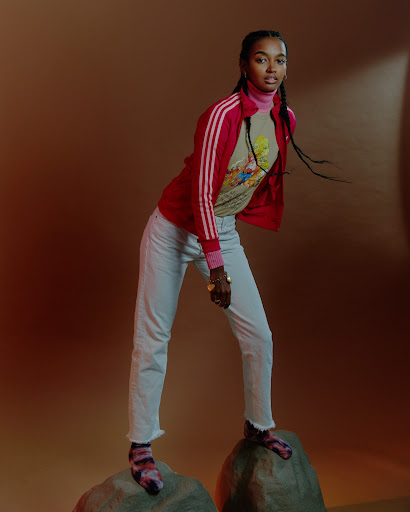 Woman in red zip jacket and white jeans standing on rocks against a brown gradient background.