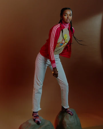Woman in red zip jacket and white jeans standing on rocks against a brown gradient background.