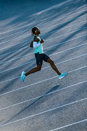 A Black adult male runner with dreadlocks and athletic wear in mid-stride on a grey outdoor running track under natural lighting.