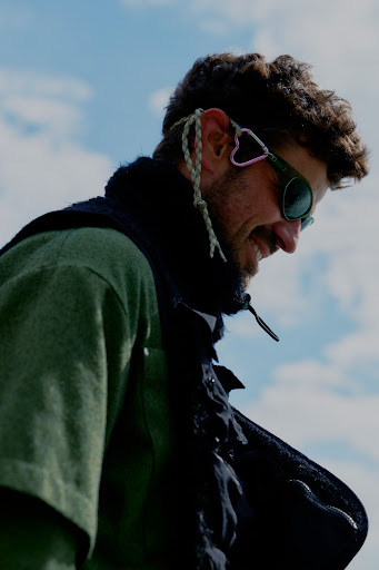 A man wearing sunglasses and a green jacket stands outdoors against a blue sky with clouds, captured in a medium shot.