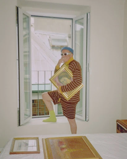 Elderly man in striped outfit poses in window holding framed painting, with additional art pieces on table in the foreground.