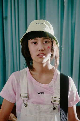Portrait of a young Asian woman wearing a Vans bucket hat and overalls, posing in front of a light blue curtain in a studio setting.
