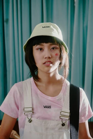 Portrait of a young Asian woman wearing a Vans bucket hat and overalls, posing in front of a light blue curtain in a studio setting.
