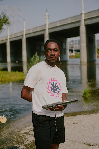 Young man stands by a river holding a keyboard, wearing a Vans t-shirt, with a bridge in the background.