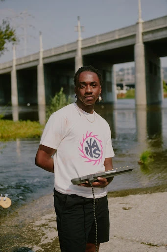 Young man stands by a river holding a keyboard, wearing a Vans t-shirt, with a bridge in the background.