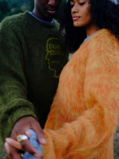 A young couple outdoors, wearing sweaters in olive and orange hues, stand together holding hands in a natural setting with soft lighting.