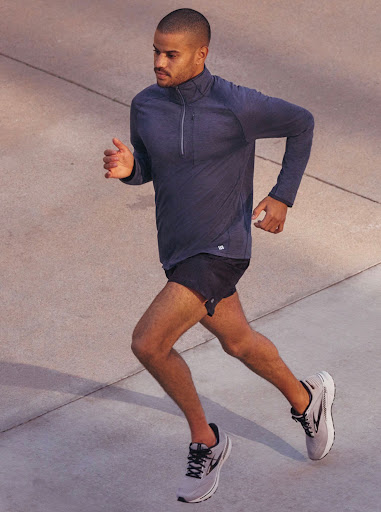 Man in athletic wear runs outdoors on concrete in eye-level shot. He wears grey running shoes, black shorts, and a long-sleeved blue shirt.