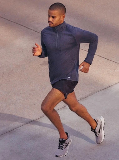 Man in athletic wear runs outdoors on concrete in eye-level shot. He wears grey running shoes, black shorts, and a long-sleeved blue shirt.