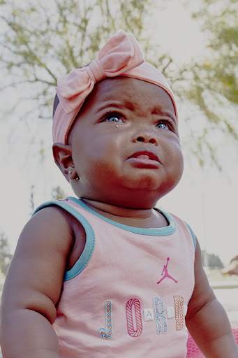 Close-up of a baby with a pink bow headband and tank top, looking up with a tear on her cheek in an outdoor setting.