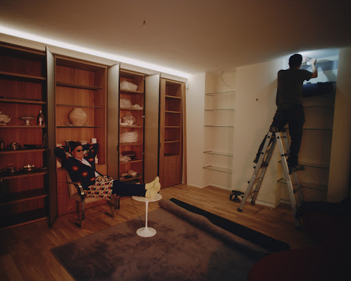 In an indoor shot, a man relaxes in a chair next to a table while another man works on shelves using a ladder in a well-lit room.