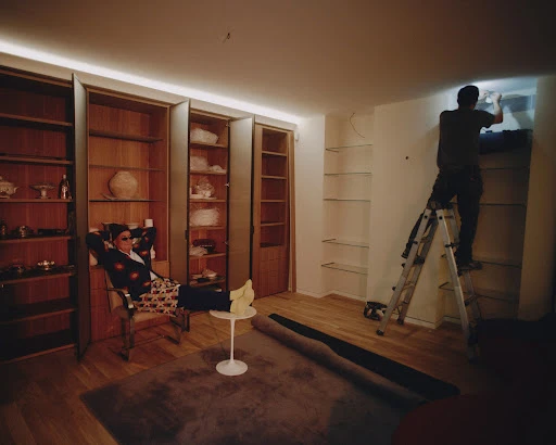 In an indoor shot, a man relaxes in a chair next to a table while another man works on shelves using a ladder in a well-lit room.