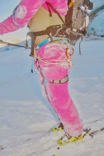 Low section of a person on skis wearing bright pink snow gear, a harness, and a backpack in a bright white snowy, outdoor setting.
