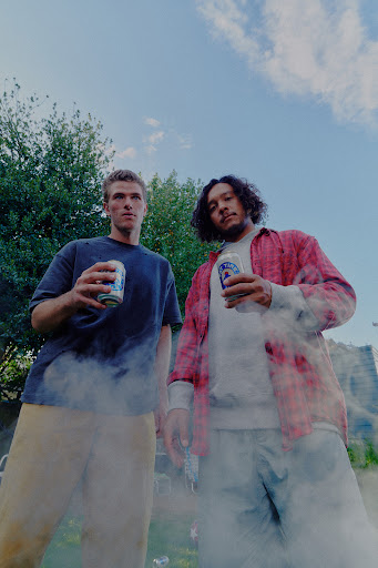 Two men stand outdoors holding canned drinks, a building and trees are visible in the background under a bright blue sky.