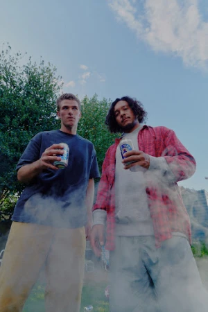 Two men stand outdoors holding canned drinks, a building and trees are visible in the background under a bright blue sky.