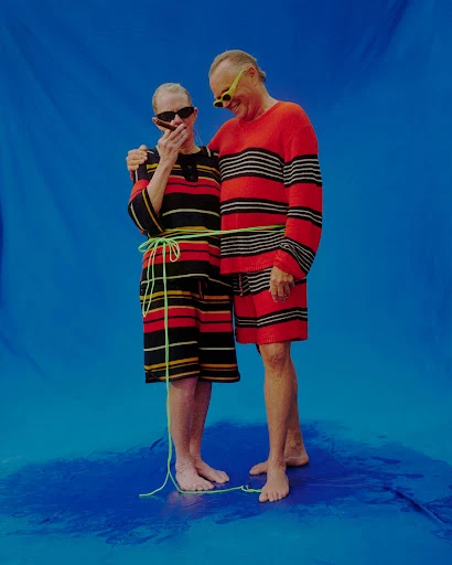 Stylish mature couple in matching red striped outfits poses on a blue backdrop, woman smoking cigar, both wearing sunglasses and tied with rope.