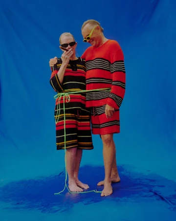Stylish mature couple in matching red striped outfits poses on a blue backdrop, woman smoking cigar, both wearing sunglasses and tied with rope.