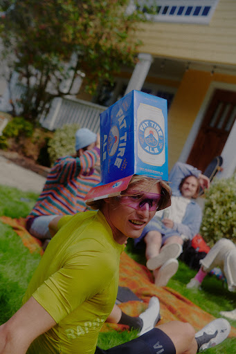 Young adult sits on a blanket in the grass, wearing a beer box as a hat, surrounded by a group relaxing outdoors on a sunny day.