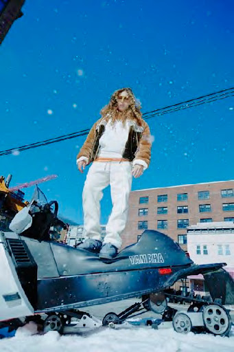 Person in faux-fur jacket poses on a black Yamaha snowmobile in snowy urban setting with blue sky during daytime.