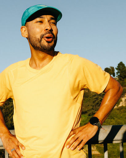 Athletic man in a yellow t-shirt and teal cap stands outdoors with hands on hips, showcasing a fitness watch and a determined expression.