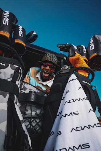 Man in patterned bucket hat smiles, surrounded by golf bags and golf clubs, bright sky visible in the background, low angle.