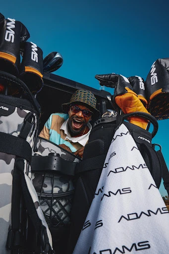 Man in patterned bucket hat smiles, surrounded by golf bags and golf clubs, bright sky visible in the background, low angle.