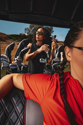 Two women in a golf cart with golf clubs. One woman faces forward, and another woman is in the golf cart with a bag of clubs.